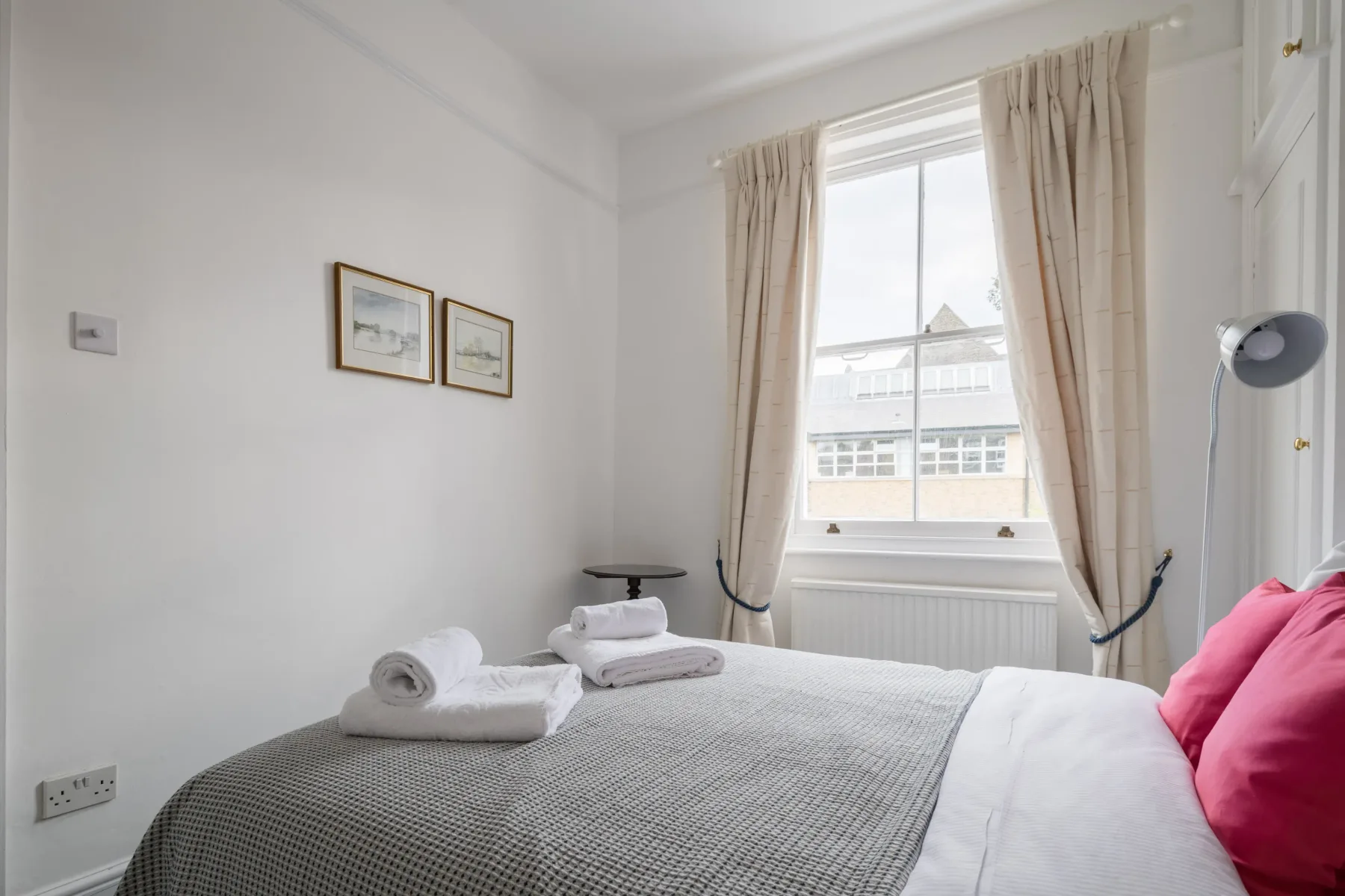 A bedroom with a neatly made bed, bright window, and pale curtains.