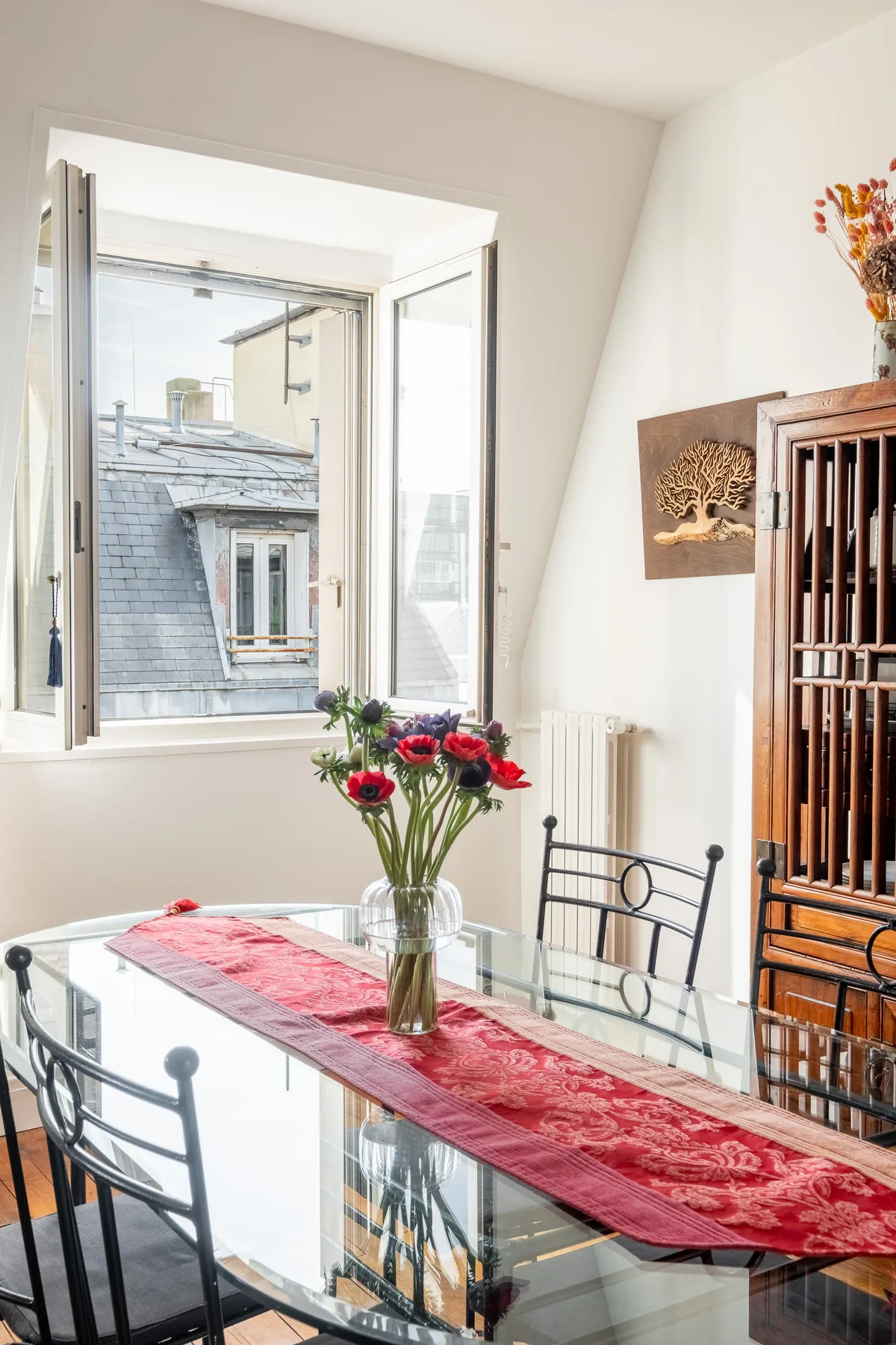 A bright dining area has a glass table set with a red runner and flowers.