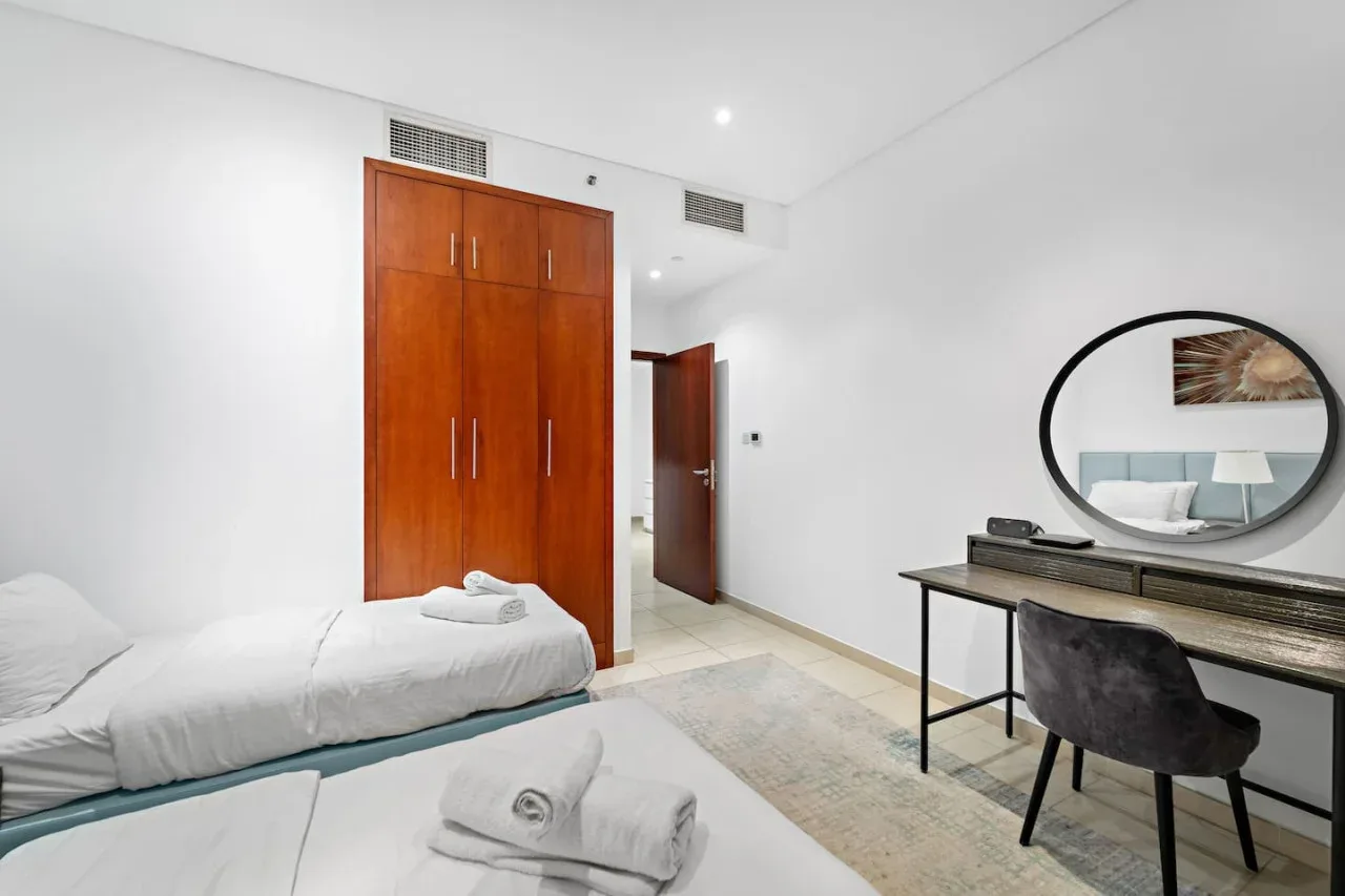 Un dormitorio luminoso con cama, armario de madera y escritorio junto a un espejo redondo.