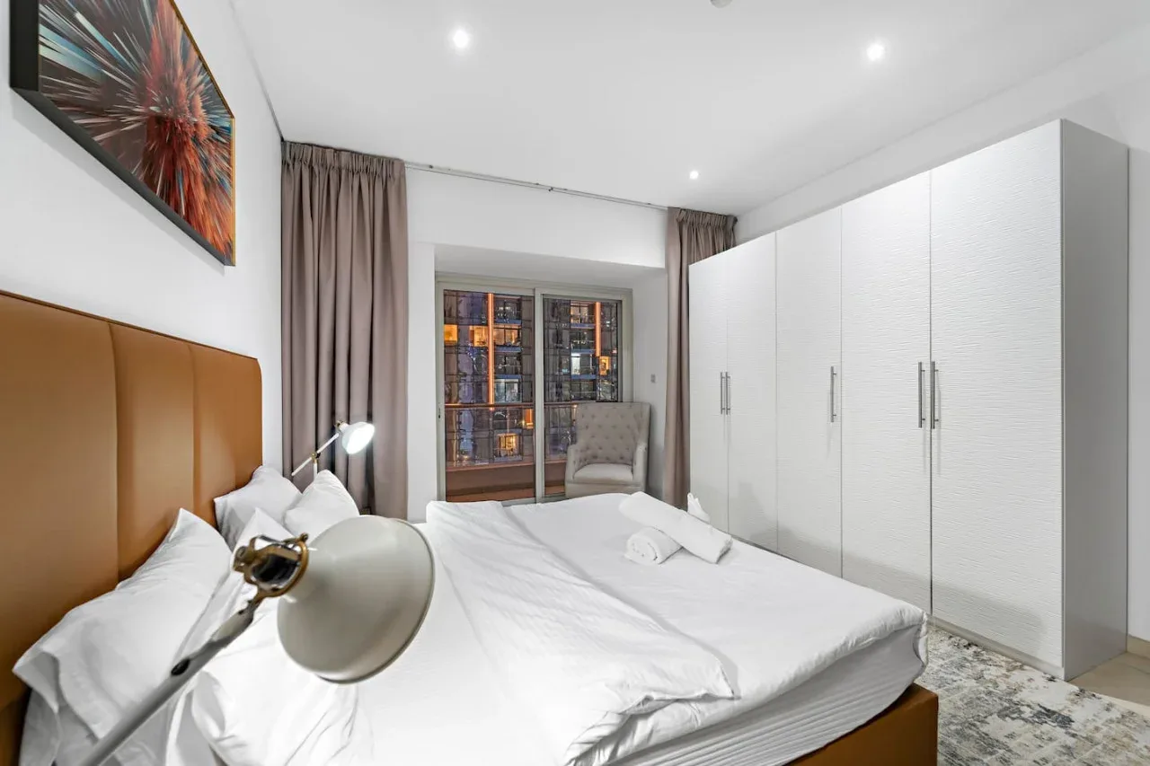 Un dormitorio luminoso con una cama blanca, armarios altos y una ventana.