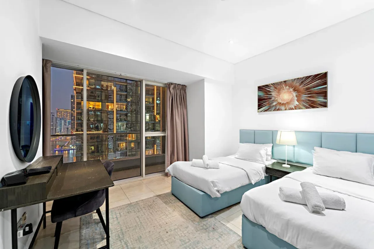 Un dormitorio luminoso con dos camas junto a una ventana grande con edificios iluminados.