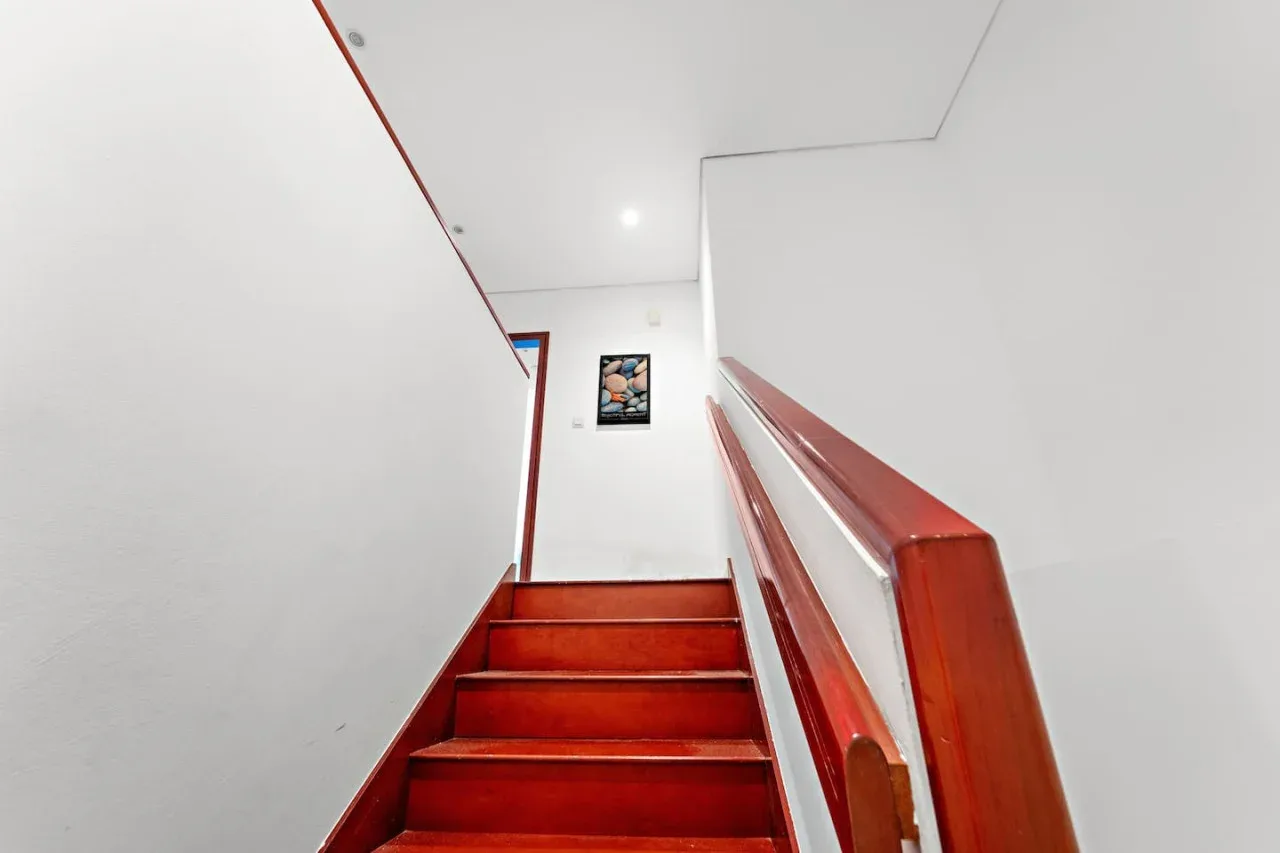 Una escalera de madera roja sube hacia un rellano blanco y luminoso con un pequeño cuadro.