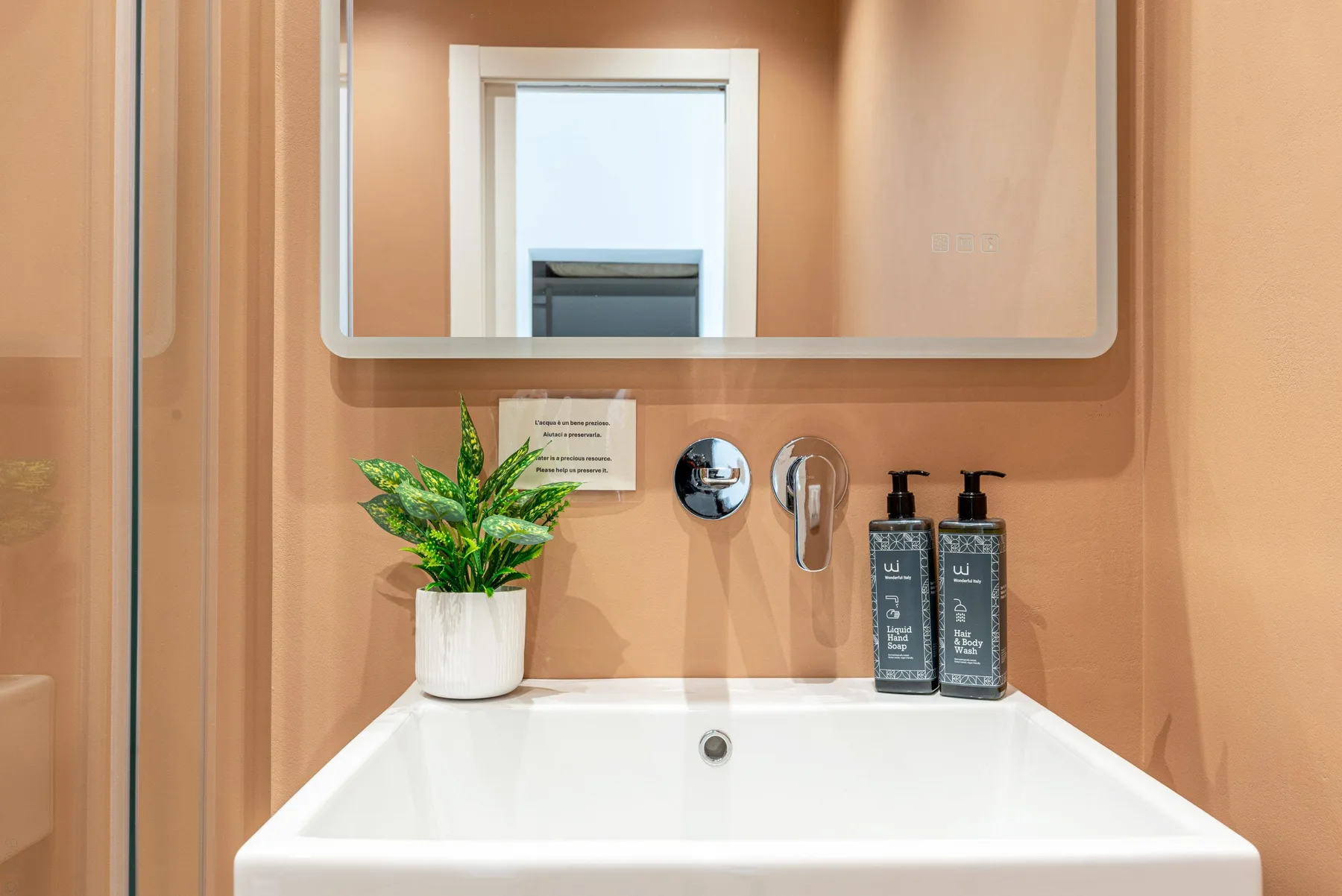 Un lavabo de baño con lavamanos blanco, espejo de pared y planta en maceta.