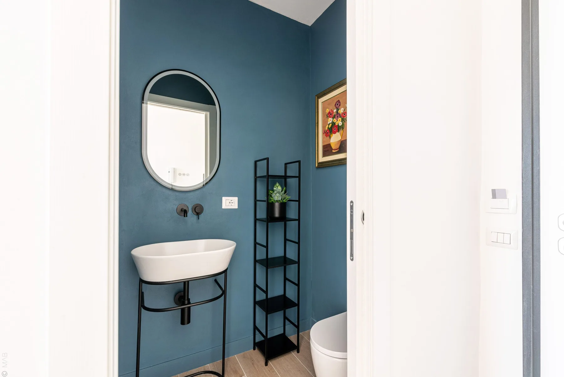 Un baño con lavabo de pared, espejo arqueado y estantería abierta.