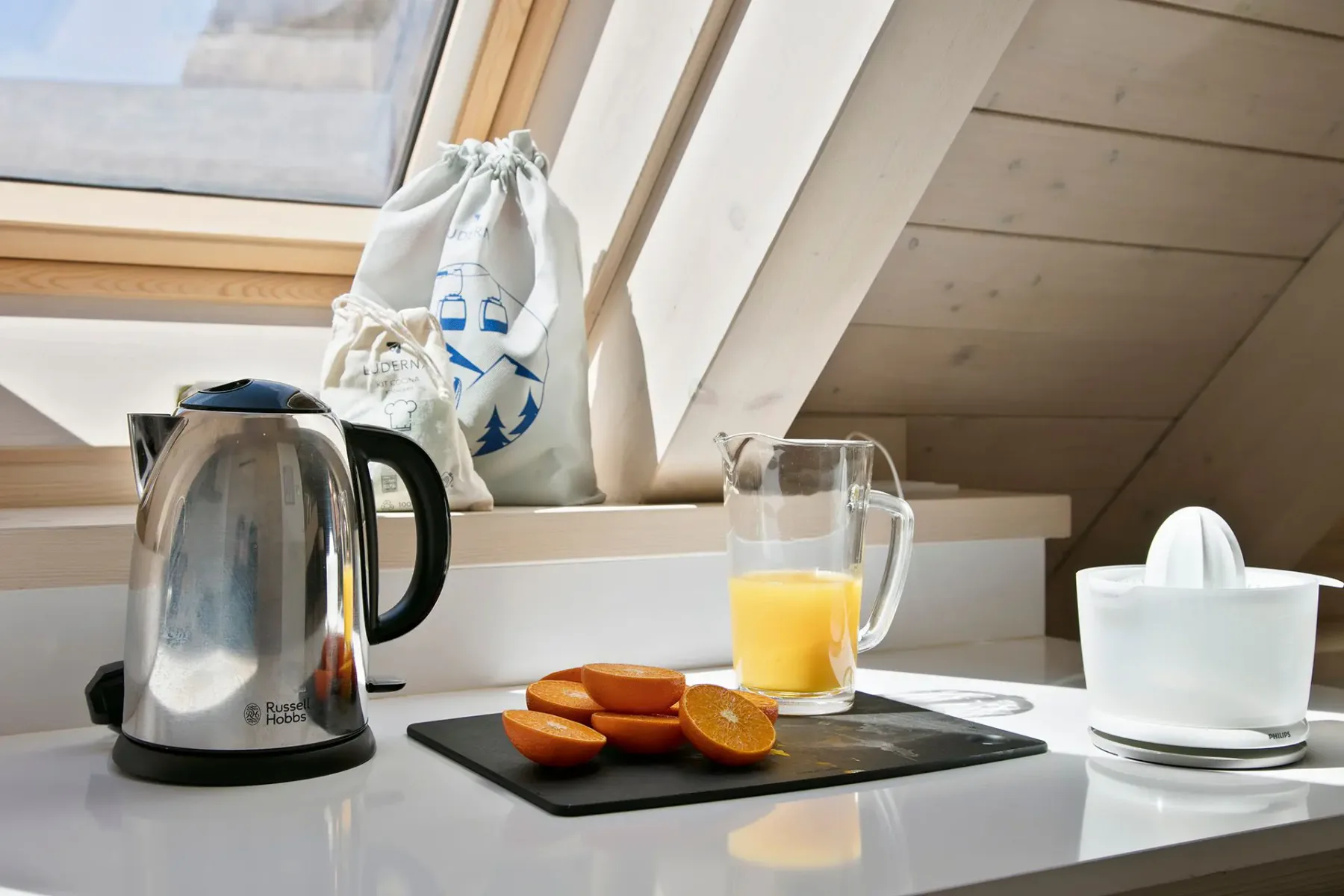 A kitchen counter with a kettle, sliced oranges on a cutting board, a jug of orange juice, and a juicer, under a attic window.