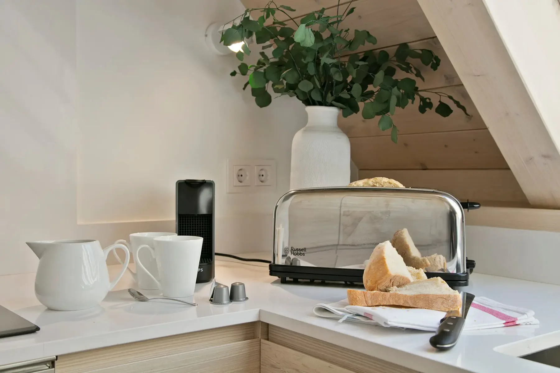 Kitchen counter with a toaster, white vase with greenery, two white mugs, and a jug. Slices of bread rest near a knife on the counter.
