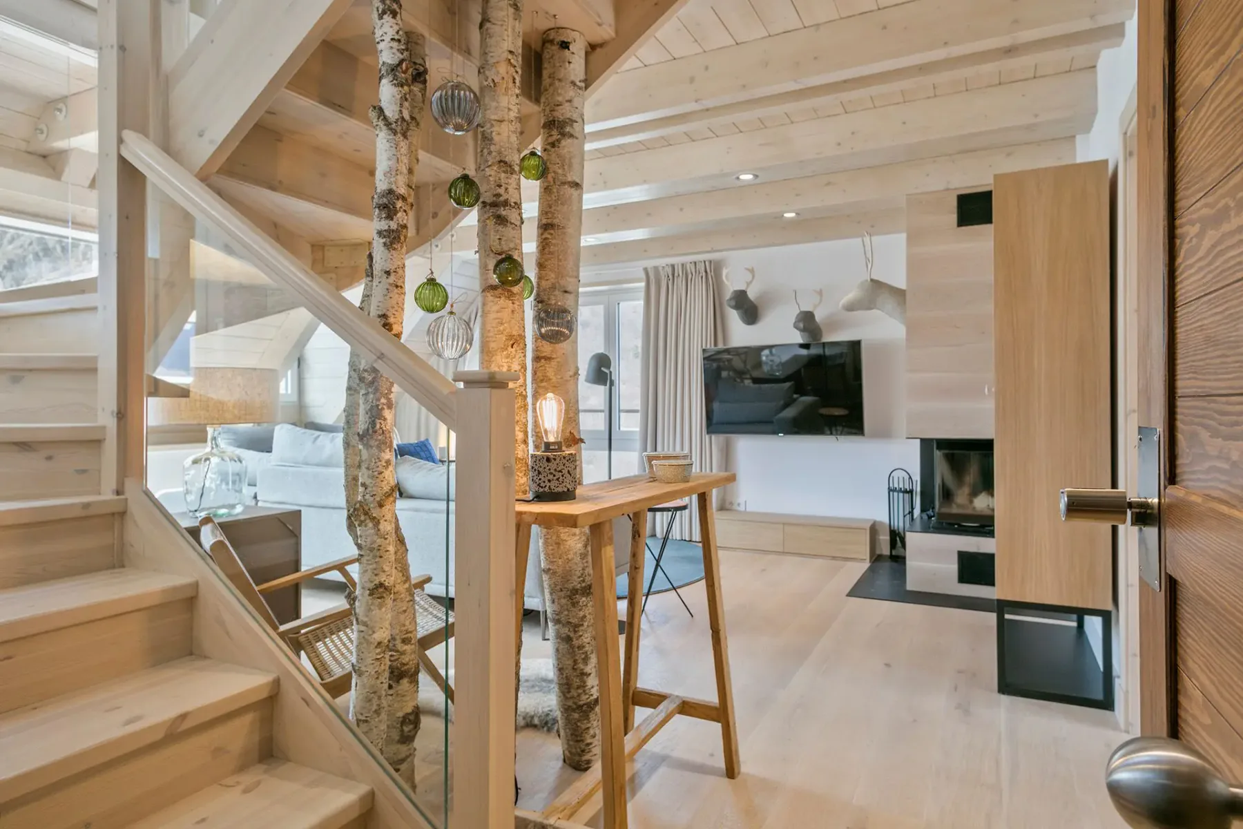 Modern living room with wooden interior, featuring a staircase, birch tree decor, a fireplace, and a mounted TV. Natural light fills the space through large windows.