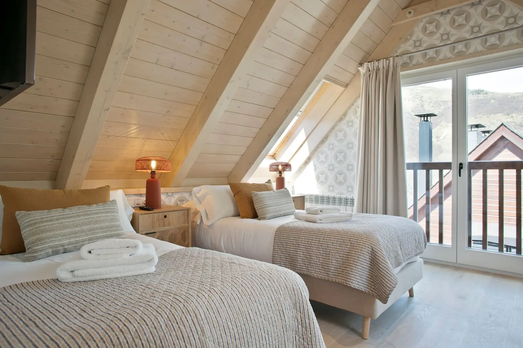 Cozy attic bedroom with two twin beds, wooden ceiling, patterned wallpaper, and large windows offering a view of the mountains.