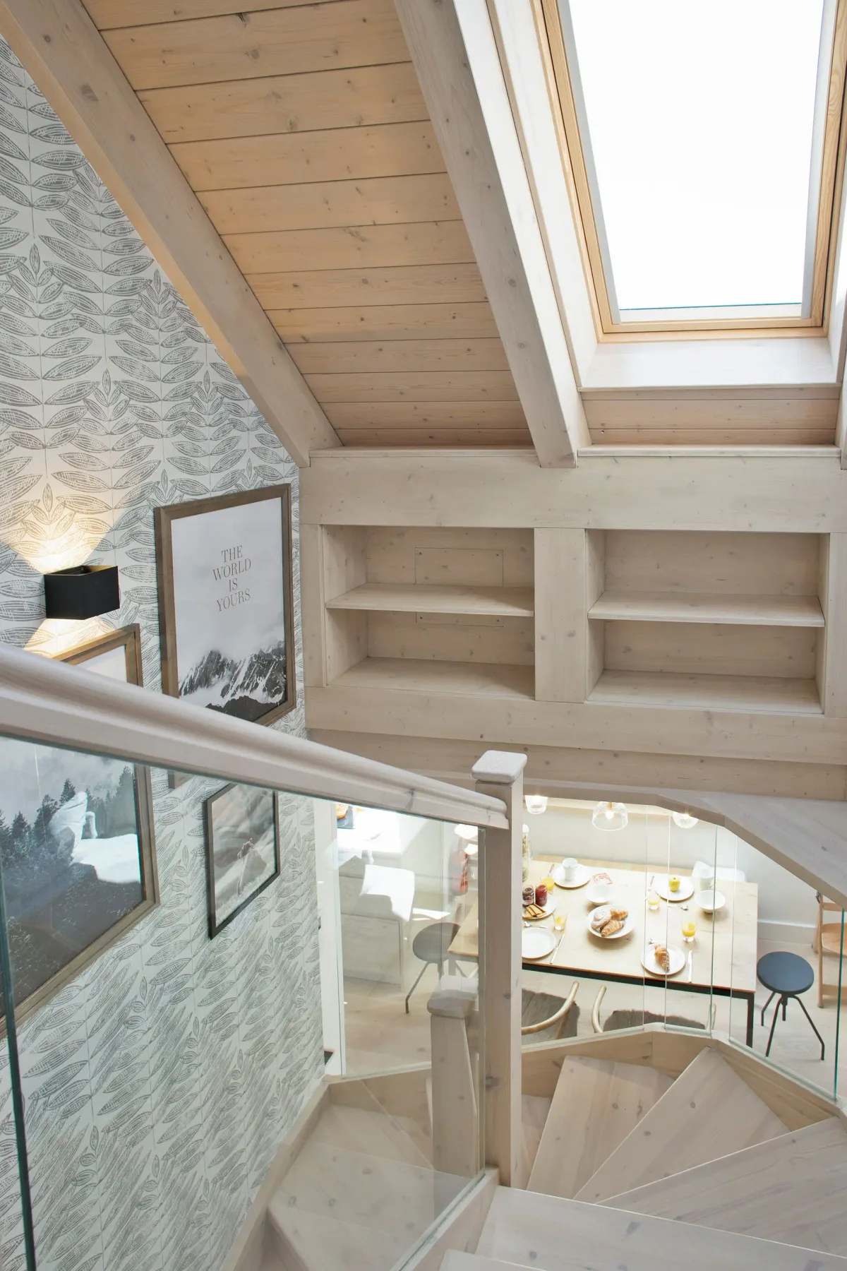 A wooden staircase with a glass railing leads to a dining area. A skylight and decorative wallpaper are visible, along with framed artwork on the walls.