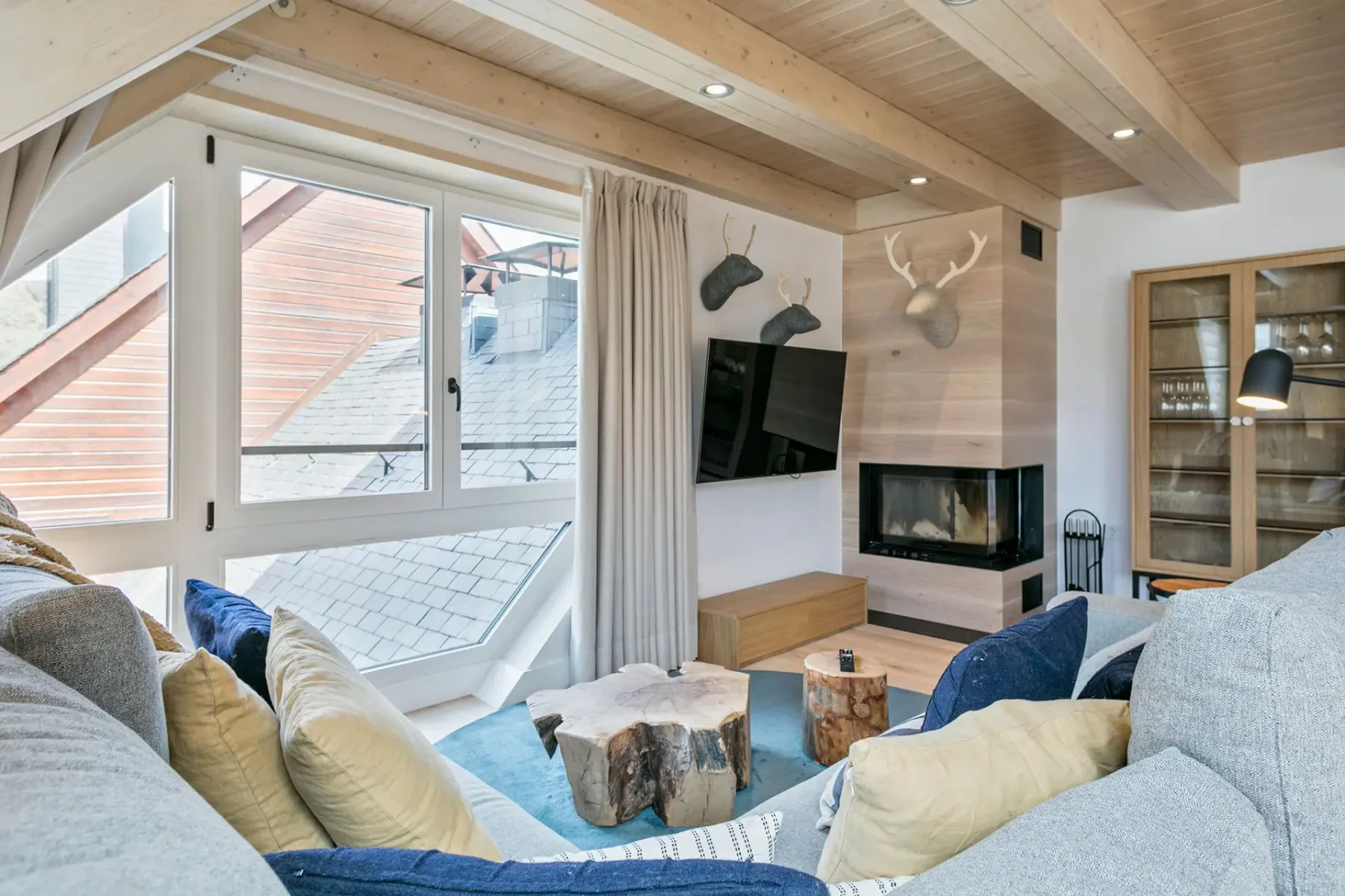 A cozy living room with a large window, wooden ceiling, sectional sofa, rustic coffee table, mounted animal heads, TV, and fireplace.