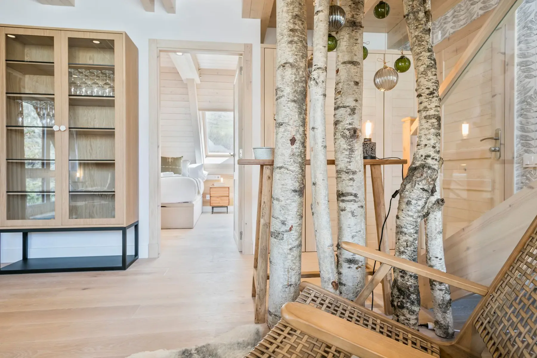 Interior of a modern wooden house with birch trees as decor, wicker chairs, and a view into a bedroom.