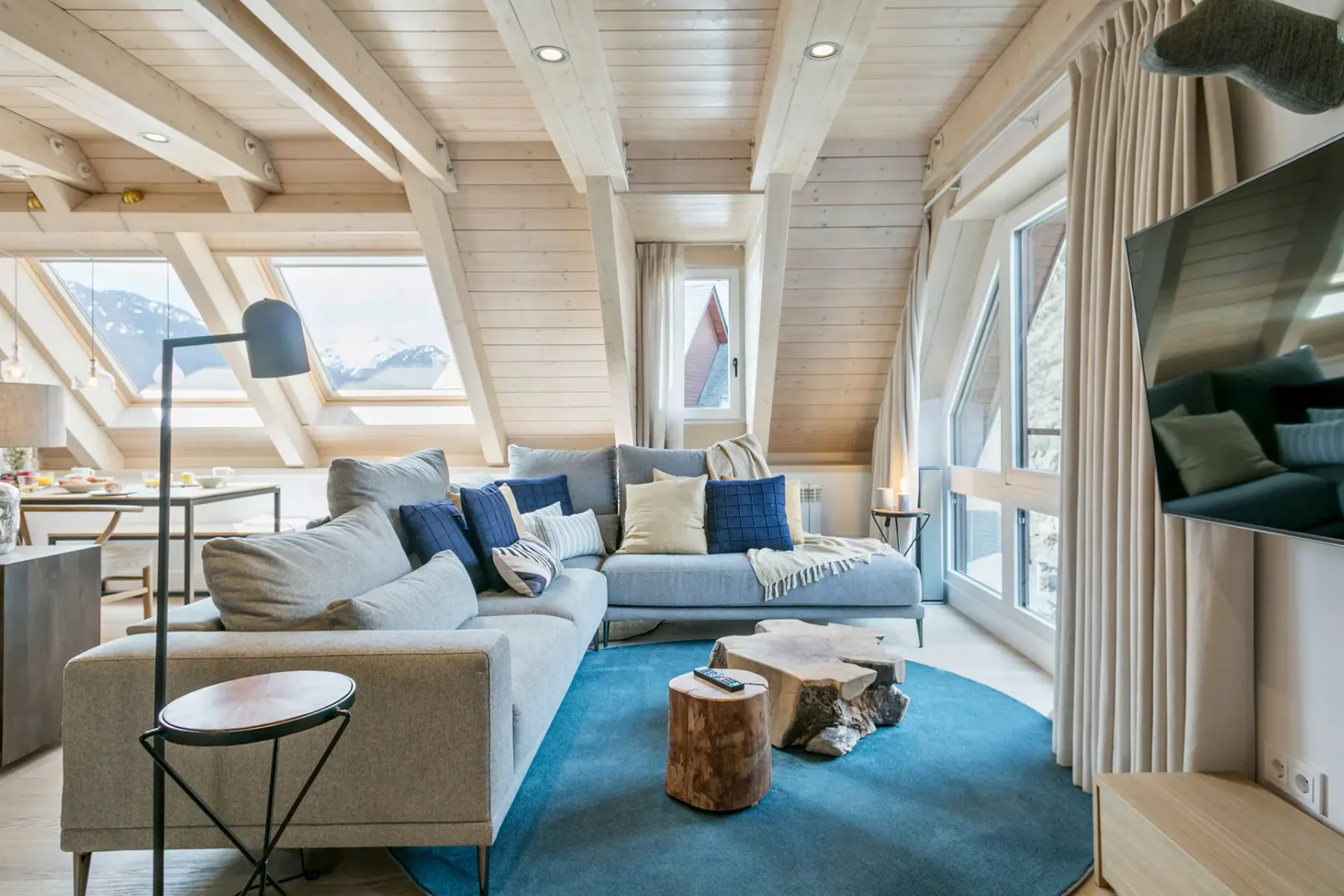 A cozy living room with a gray sectional sofa on a blue rug, wooden ceiling beams, a wood coffee table, and large windows showing a mountain view. A dining area is visible in the background.
