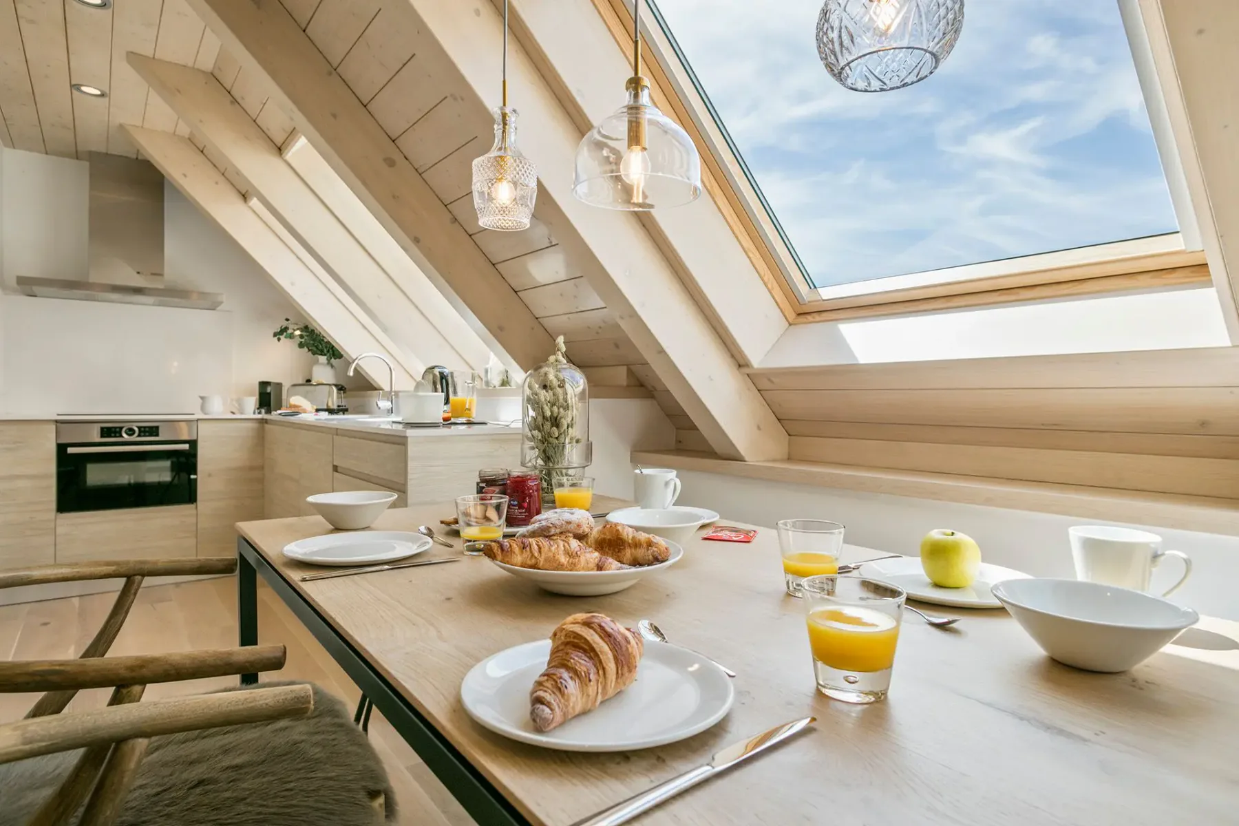 Modern kitchen with wood accents, skylight, and breakfast setup. Table includes plates, croissants, orange juice, an apple, and coffee cups. Natural light illuminates the space.