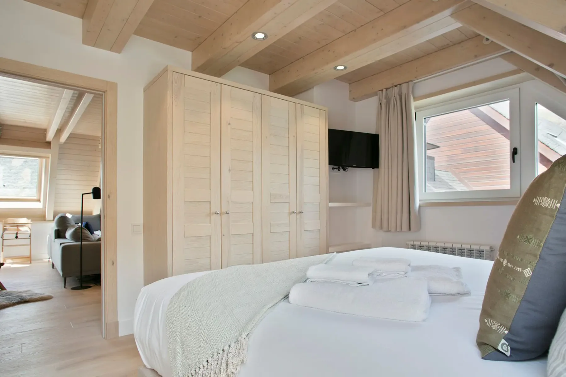 Cozy bedroom with a large bed, wooden wardrobe, and a wall-mounted TV. Natural light filters through a window. Visible entry to a living space with a couch.