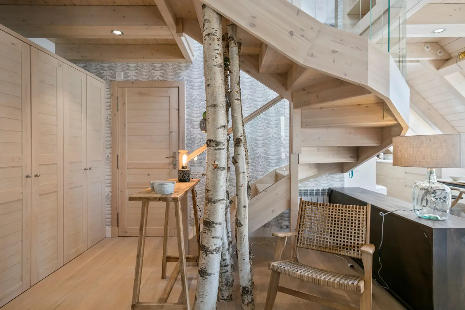 A bright wood-paneled interior with a staircase, birch trunks, and a small dining table.