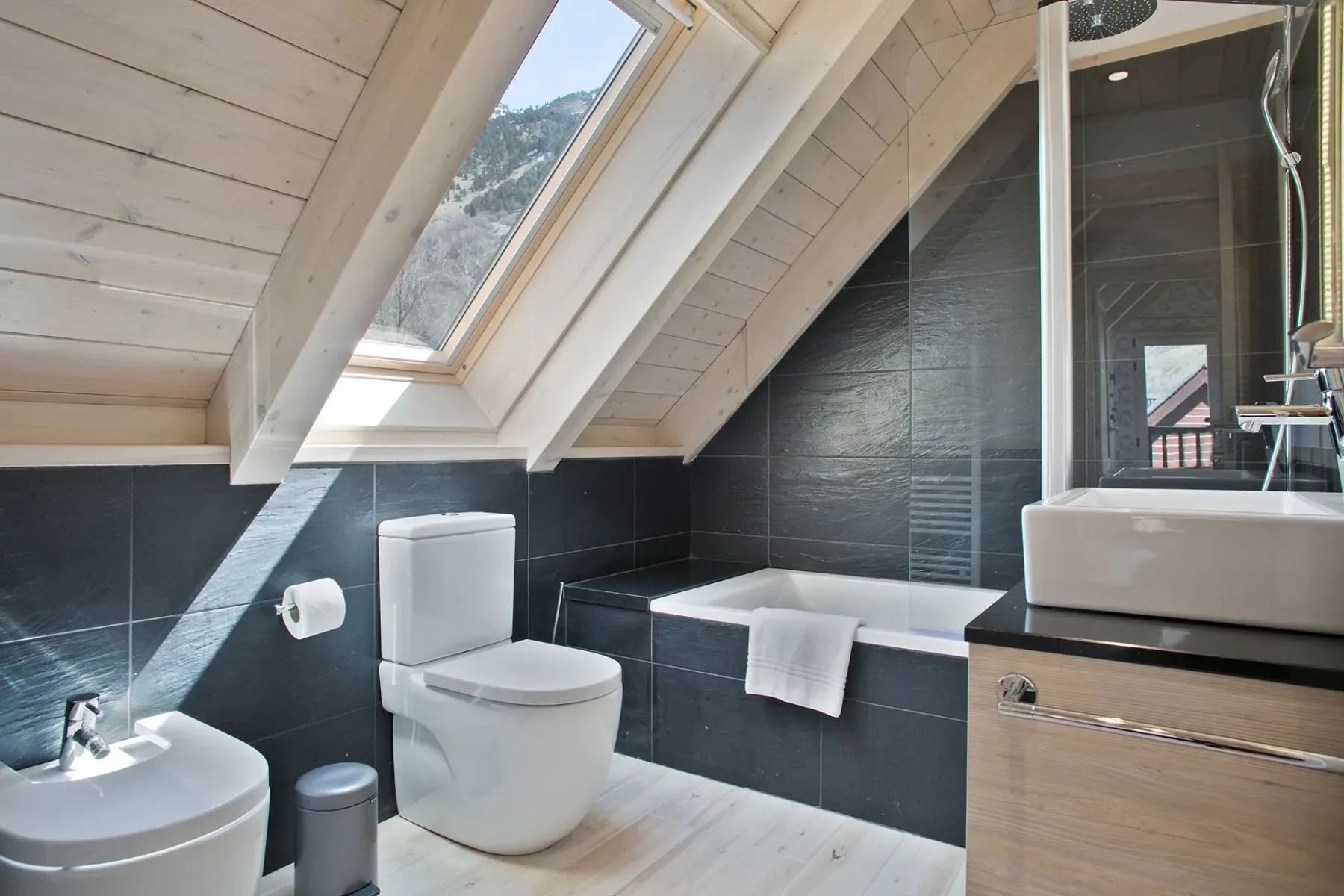Modern attic bathroom with black tiles, a skylight, toilet, bidet, and a bathtub. Light wood paneling and flooring add contrast to the space.