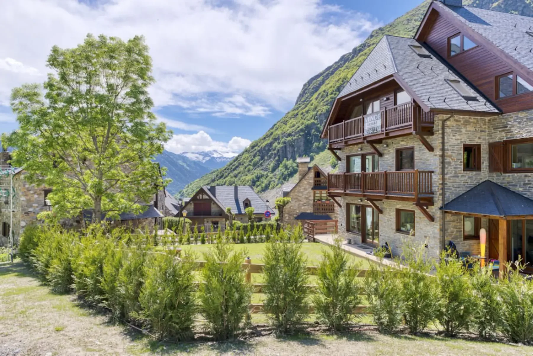 Mountain chalet with stone and wood exterior, surrounded by greenery and small hedges, set against a backdrop of hills and a blue sky.