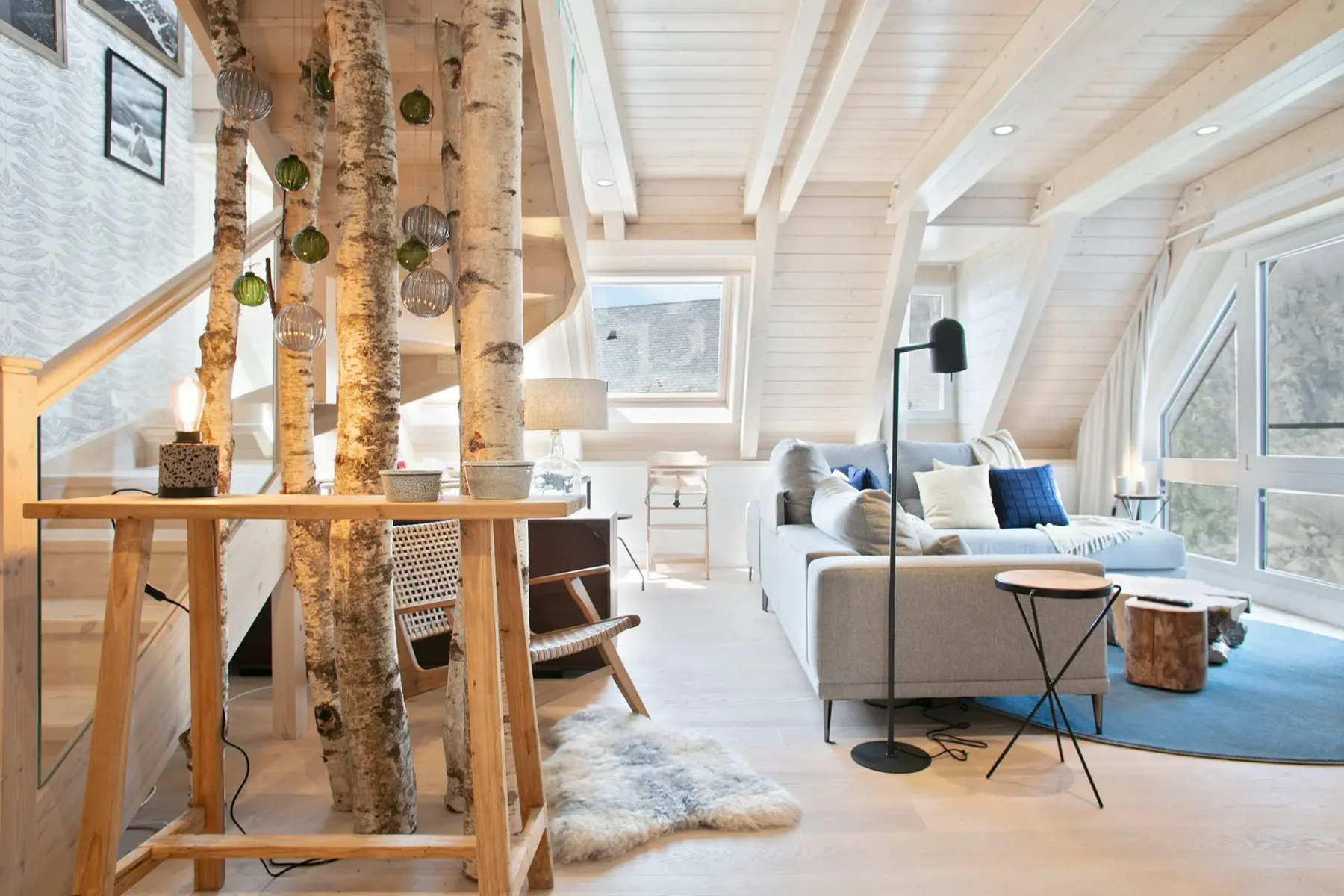 Modern living room with large windows, wood beams, a gray sofa, and birch tree decor. Natural light highlights a cozy, minimalist design with neutral tones and a mix of textures.