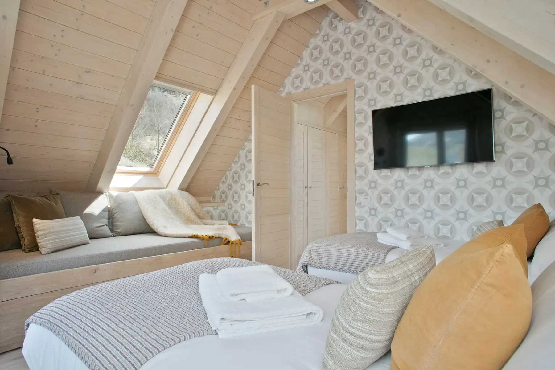 A cozy attic bedroom with wooden paneling, twin beds, a skylight, patterned wallpaper, a wall-mounted TV, and a padded bench with cushions.