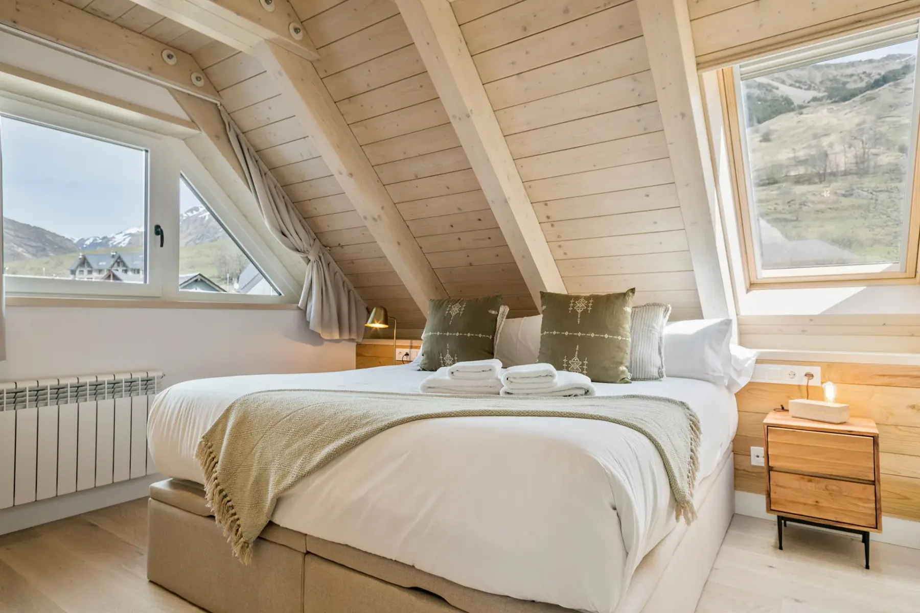 Cozy attic bedroom with a large bed, wooden walls and ceiling, two windows showing a mountain view, and bedside tables with lamps.