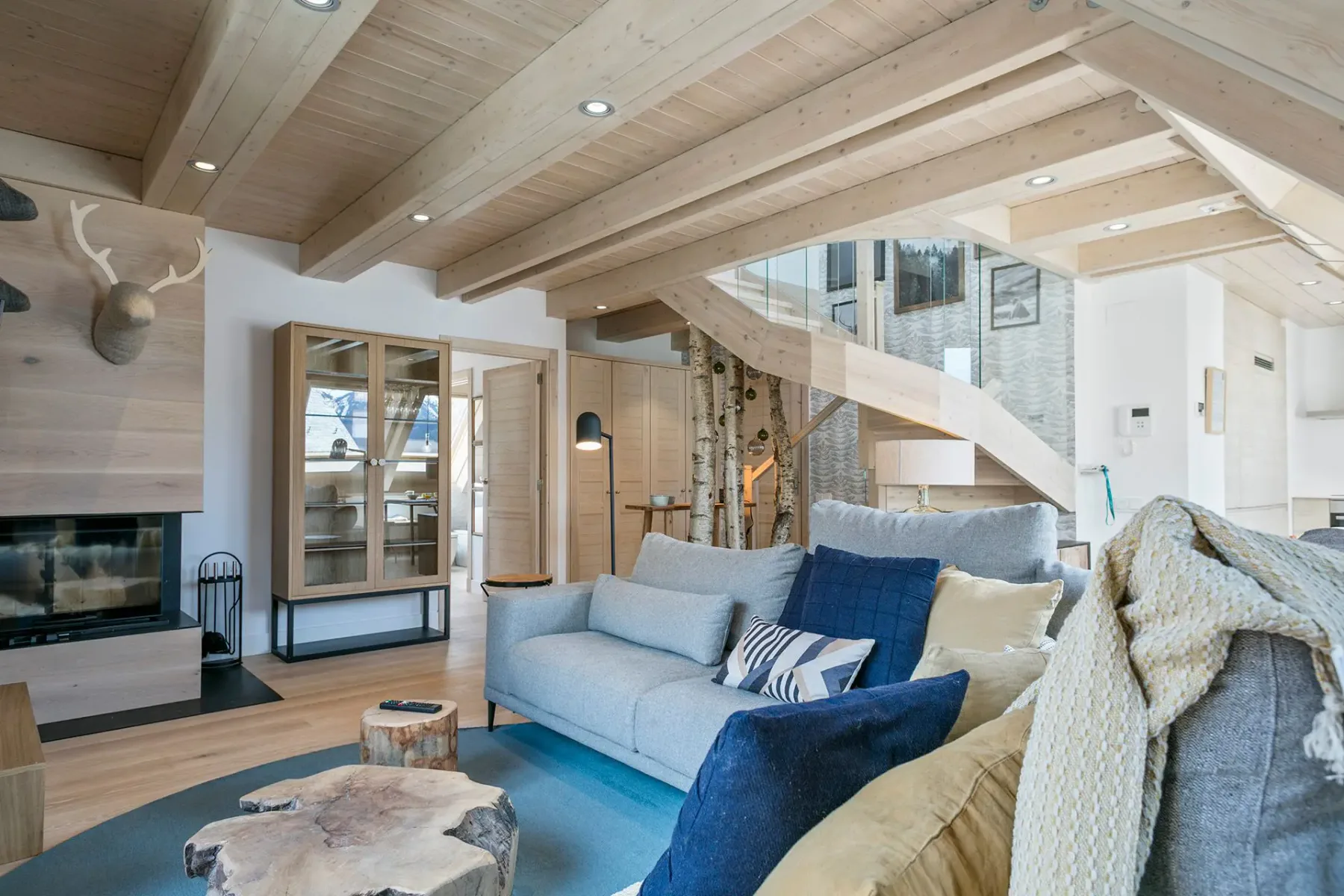 Cozy modern living room with wooden ceiling, light gray sofa, multicolored cushions, deer wall mount, and a unique wooden coffee table. Stairs lead to an upper level.