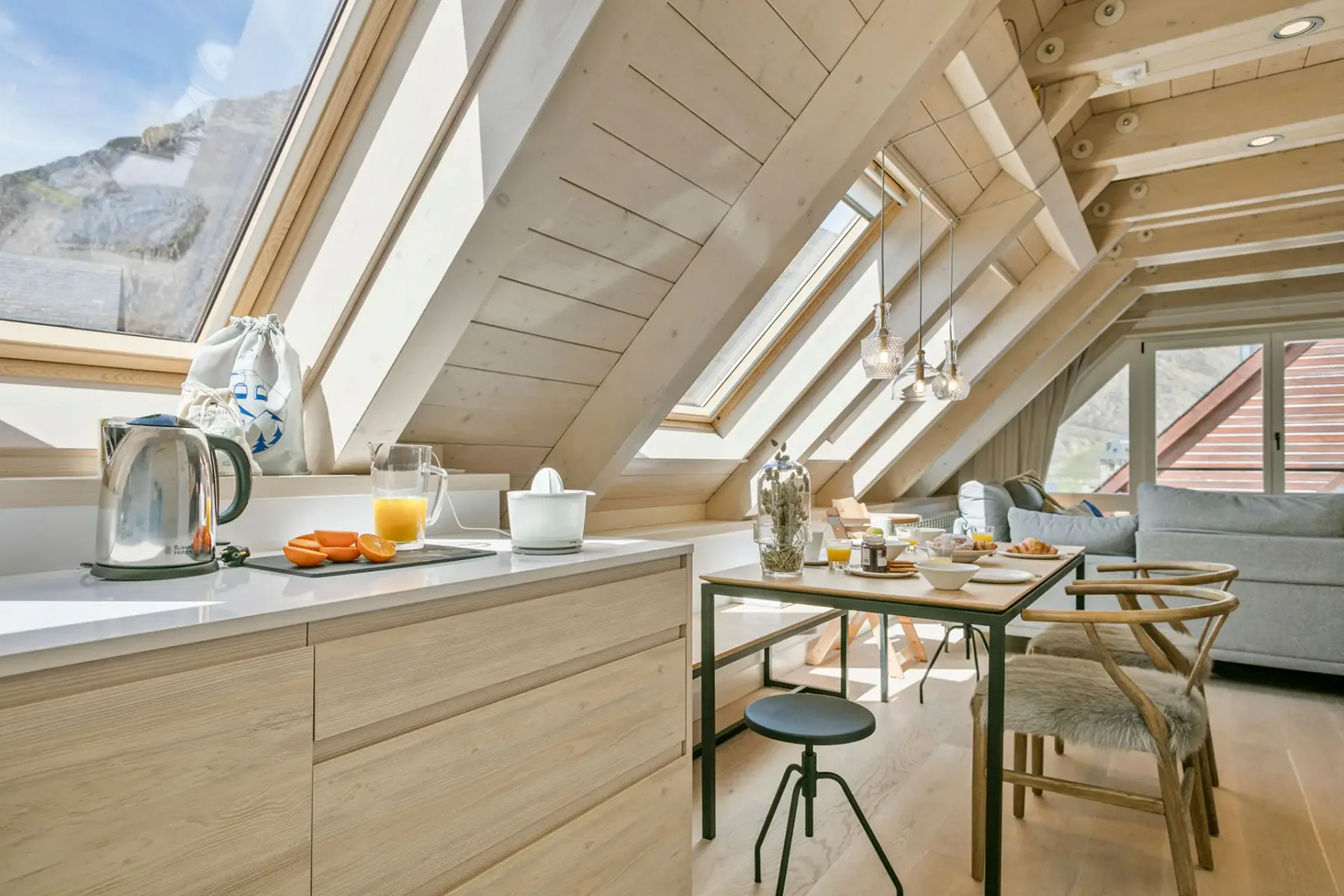 A bright attic kitchen with skylights, wooden cabinets, a small dining table set for breakfast, chairs, and a sofa in the background.