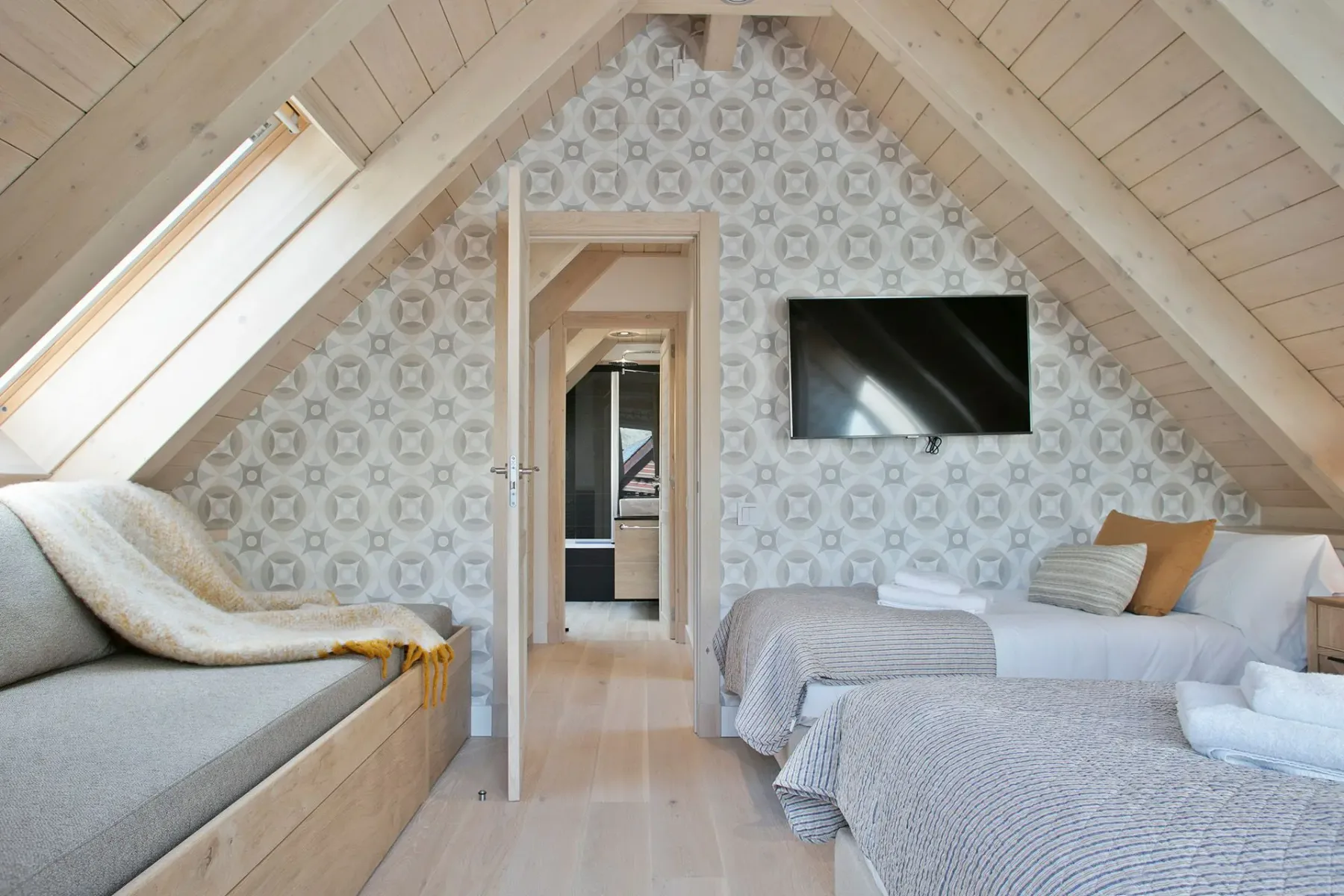 Attic bedroom with two beds, wall-mounted TV, and patterned wallpaper. Open door reveals a connecting hallway. Natural light from skylight illuminates the wooden interior.