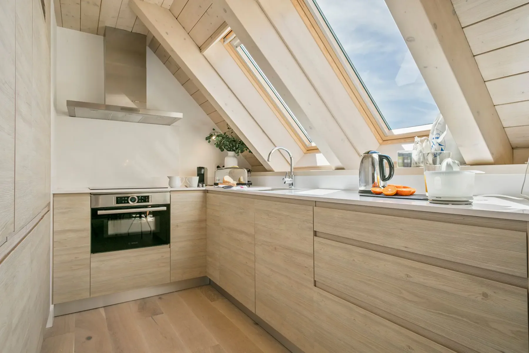 A modern kitchen with light wood cabinets, a built-in oven, and large skylights. A kettle and cutting board with oranges are on the countertop.