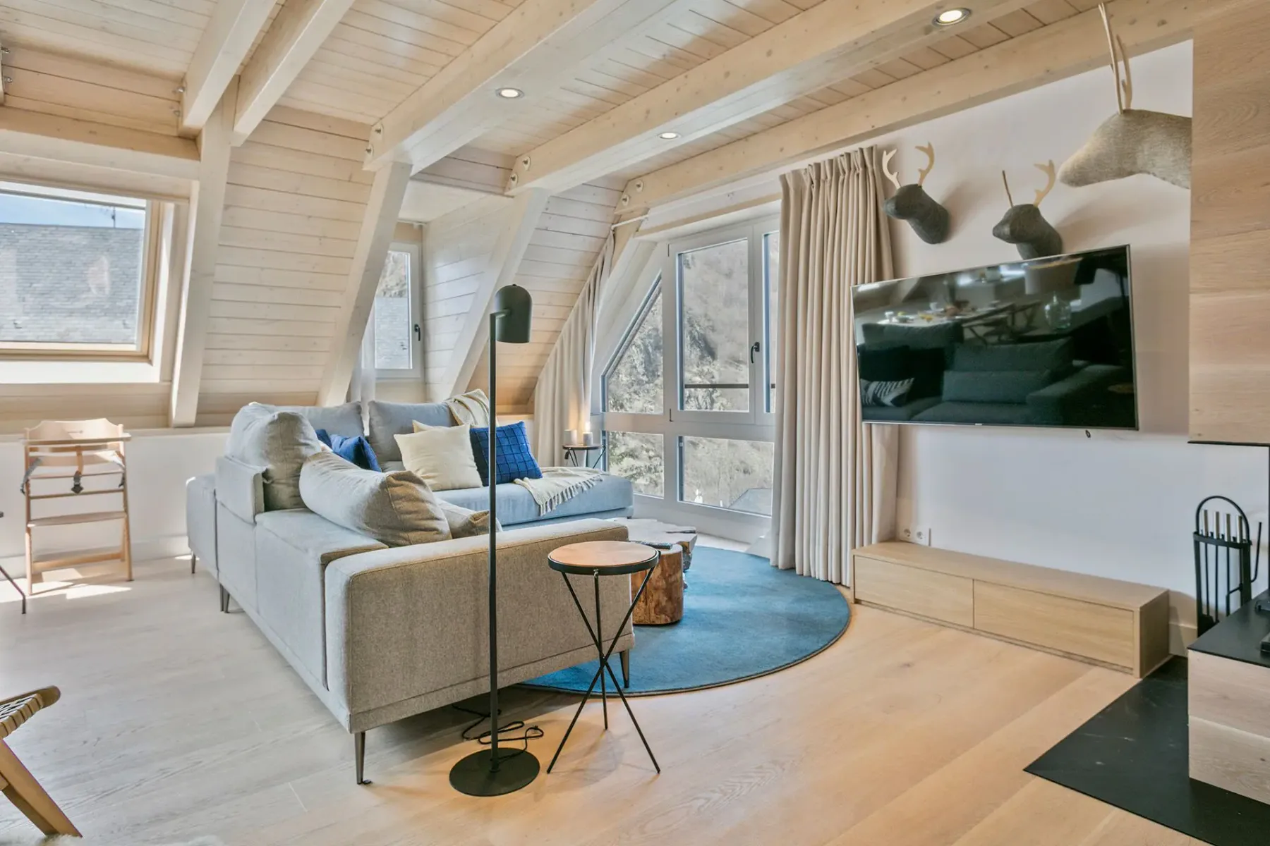 A modern living room with a gray sofa, blue cushions, a round rug, a floor lamp, a TV on the wall, and decorative wall-mounted deer heads. The room has wooden walls and large windows.
