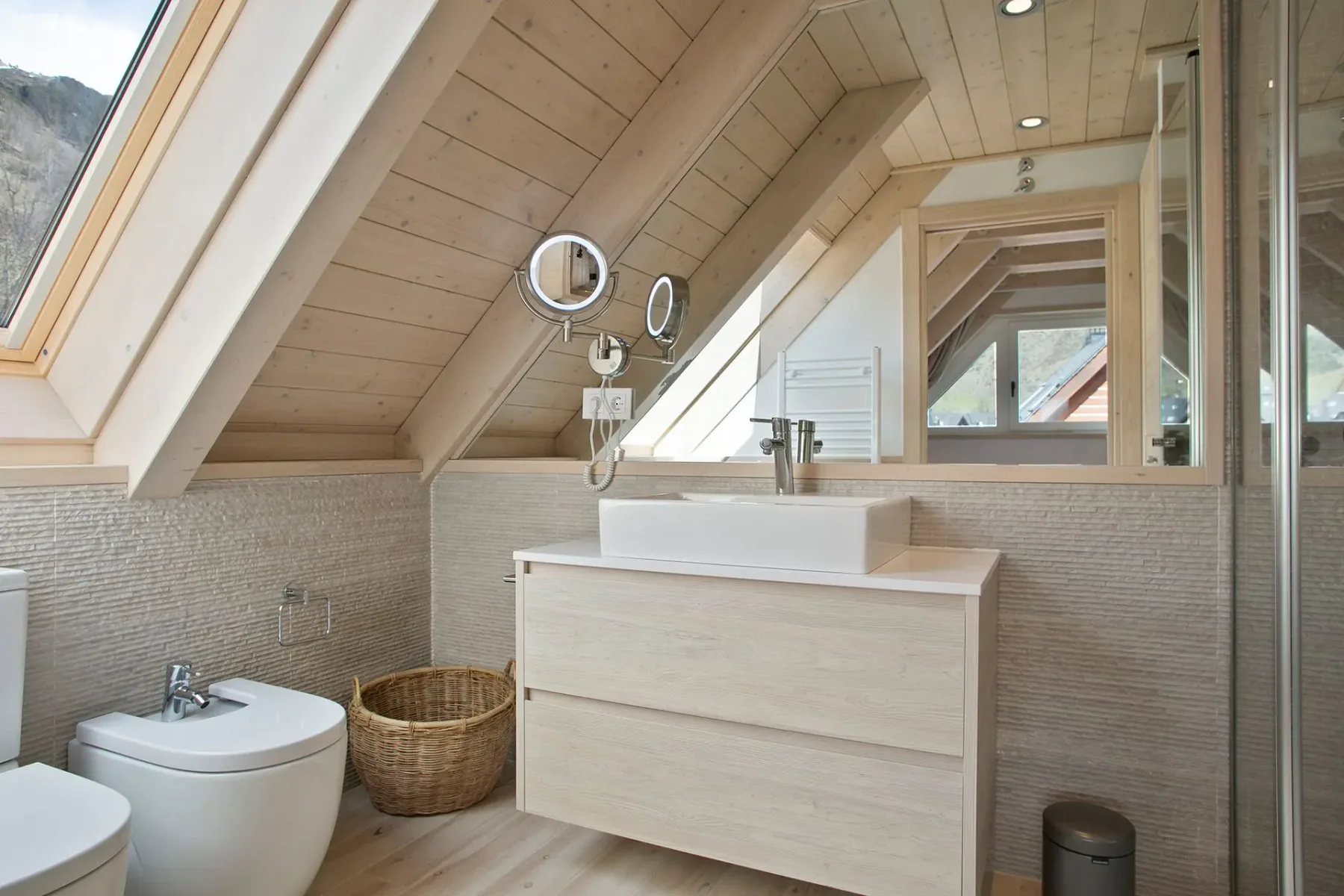 A modern attic bathroom with slanted wooden ceiling, a window, a vanity with rectangular sink, a round mirror, and a wicker basket.