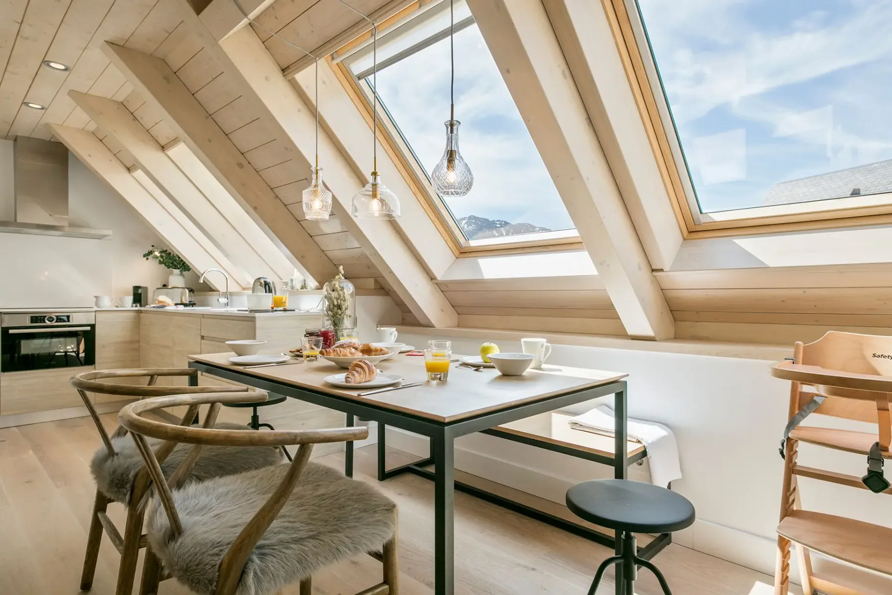 A modern kitchen with wooden beams, a dining table set for breakfast, and large skylight windows providing natural light.