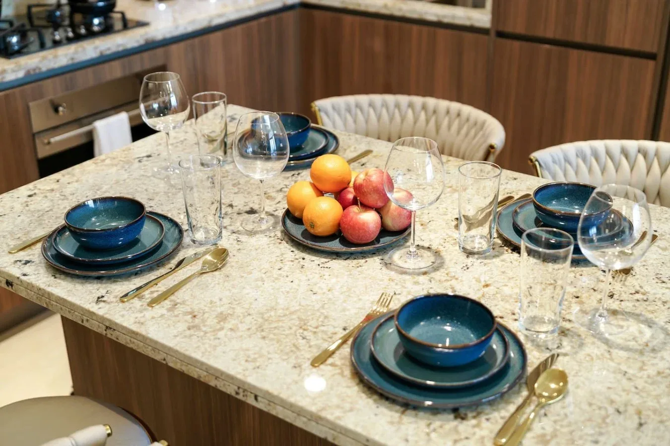 Una isla de cocina está preparada con platos, cristalería y un cuenco de fruta.
