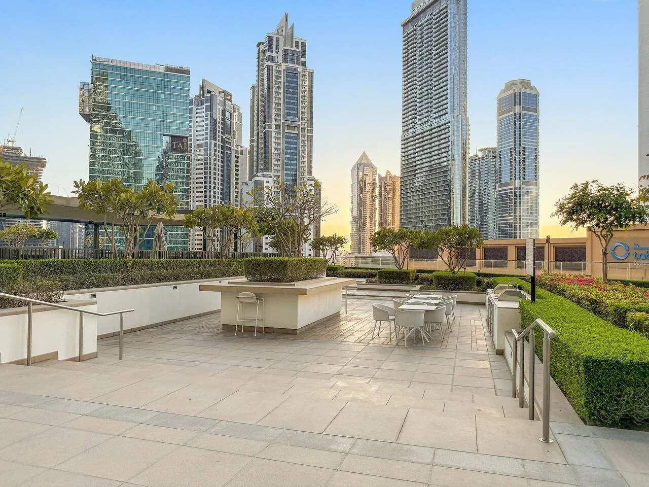 Una terraza exterior pavimentada con asientos y jardineras ajardinadas da a un conjunto de edificios altos.