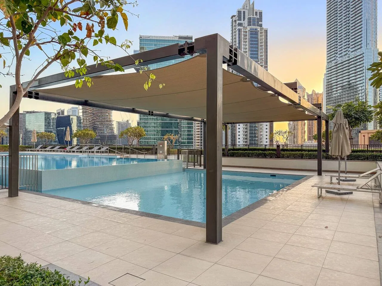Una piscina al aire libre está bajo un amplio toldo, con edificios urbanos al fondo.
