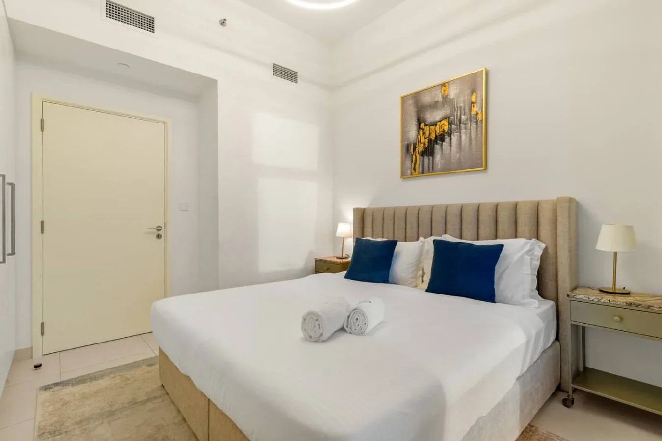 Un dormitorio luminoso con una cama grande, cojines azules y un cuadro en la pared.