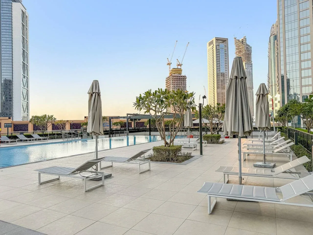 Una terraza de piscina al aire libre con tumbonas y sombrillas cerradas, rodeada de edificios altos.