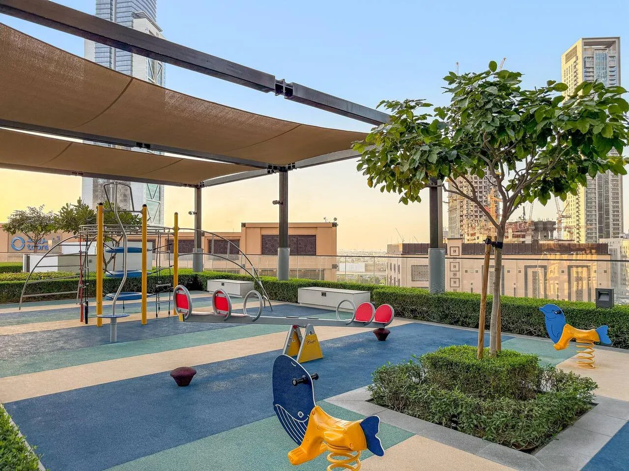 Un parque infantil al aire libre con sombra, juegos coloridos y un árbol.