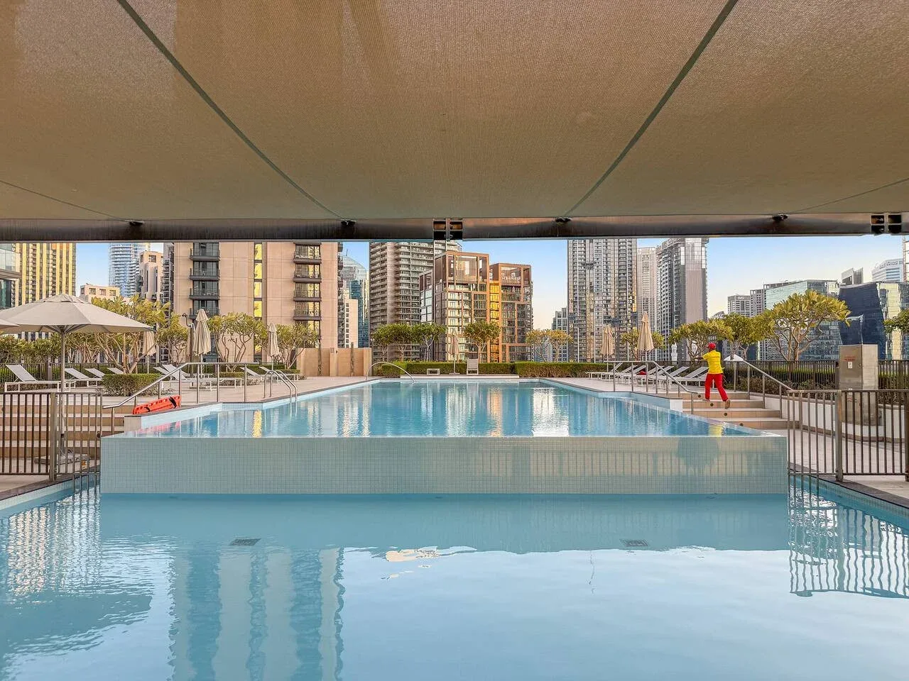 Una piscina al aire libre está bajo un toldo, con edificios urbanos al fondo.