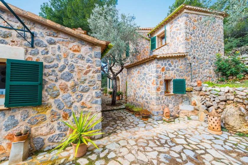 A stone courtyard between rustic buildings, with green shutters and potted plants.