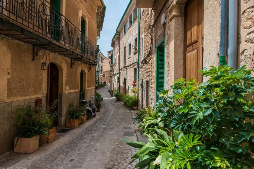 A narrow cobblestone street runs between old stone buildings and greenery.