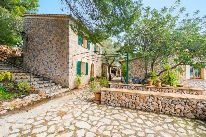 A stone house opens onto a paved courtyard with trees and potted plants.