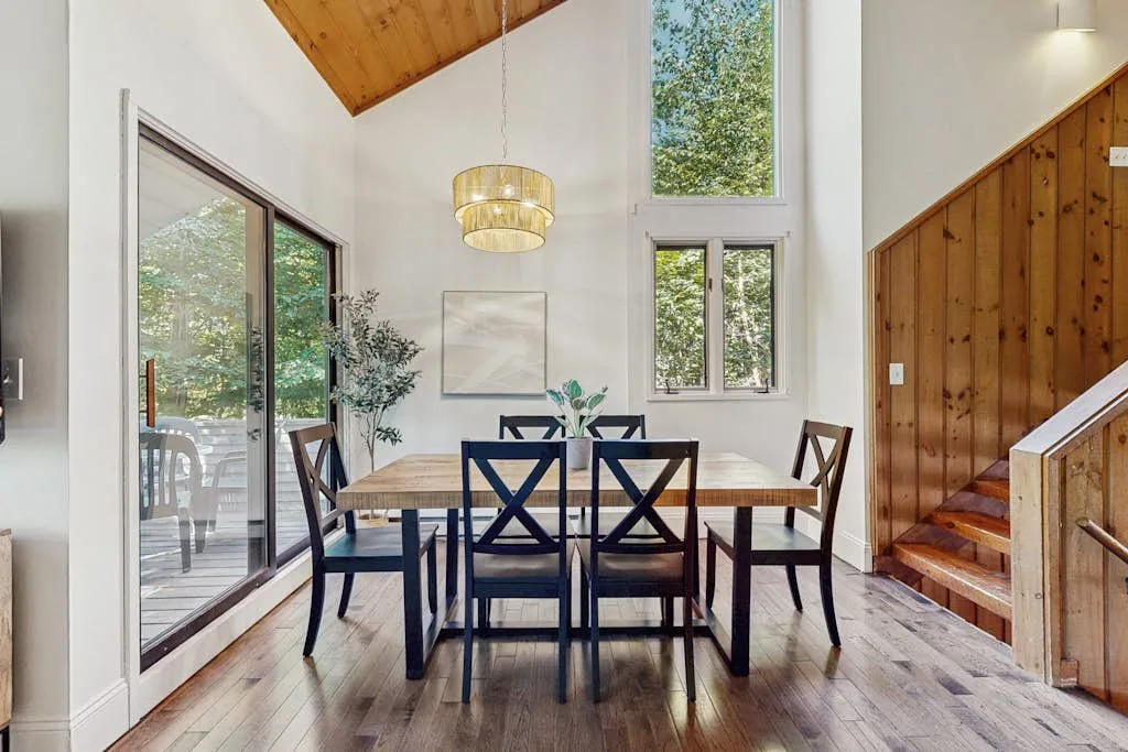 A dining area with a wooden table and chairs beside large windows.