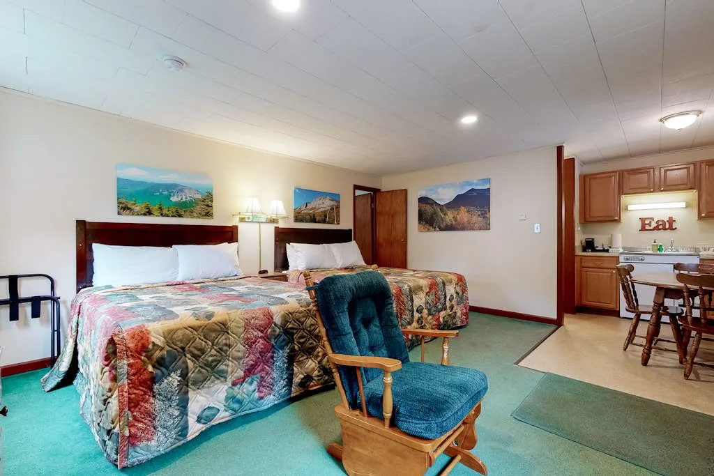 Un dormitorio con dos camas y una silla verde azulado, con una pequeña cocina al fondo.