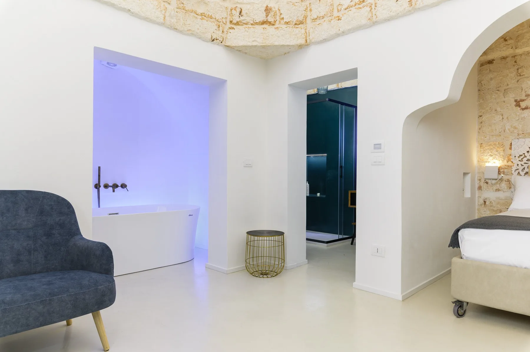 A bright bedroom with a white bed, a blue armchair, and a bathtub.
