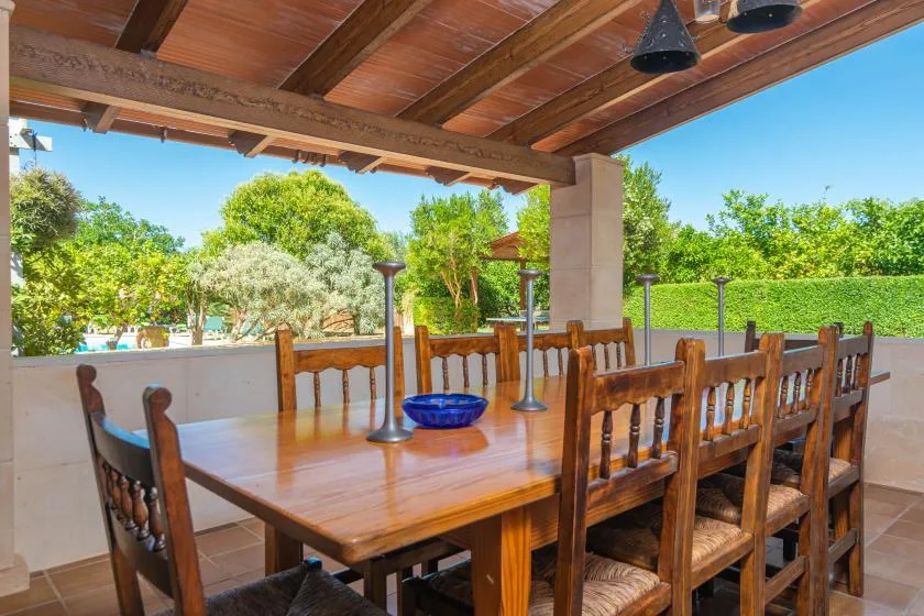A covered outdoor dining area with a long wooden table and surrounding greenery.