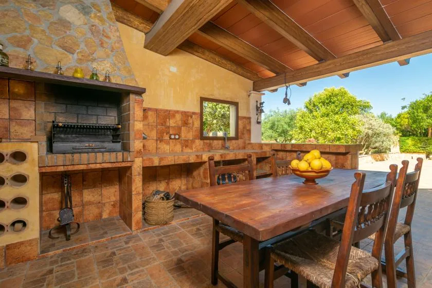 A covered outdoor dining area has a long wooden table beside a brick barbecue.