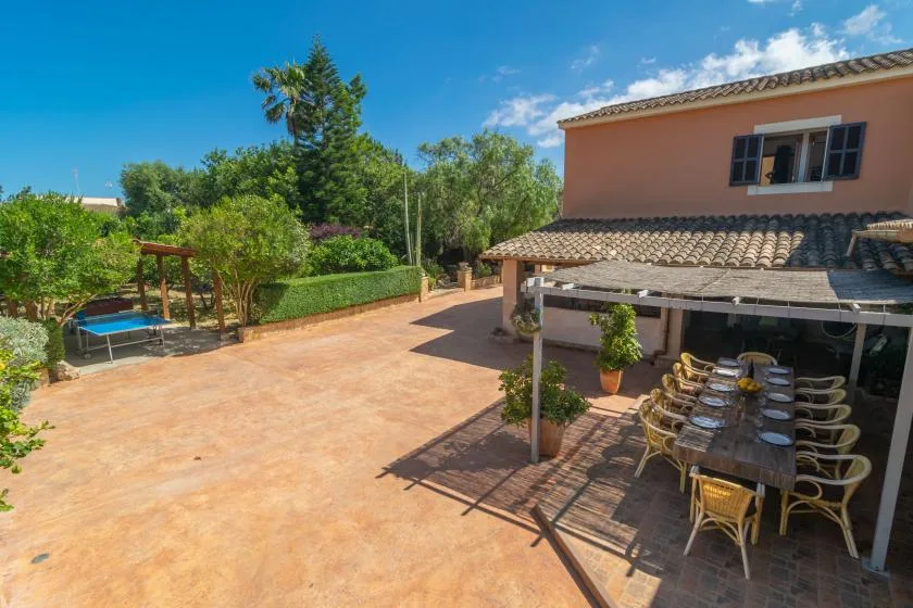 A sunny outdoor patio with a long dining table beneath a tiled canopy.