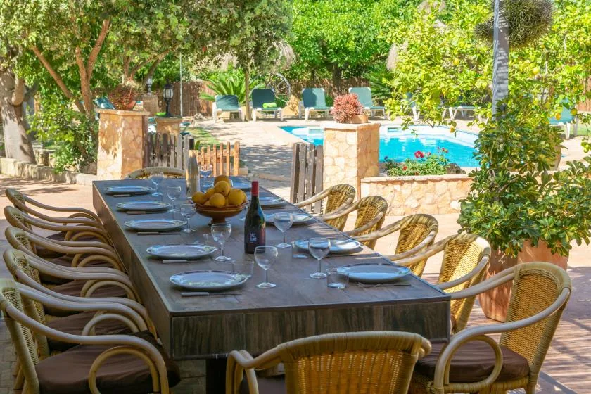 An outdoor dining table is set beside a swimming pool amid leafy garden surroundings.