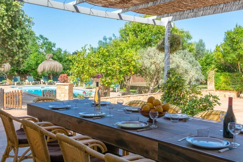 An outdoor dining table sits beneath a pergola beside a swimming pool.