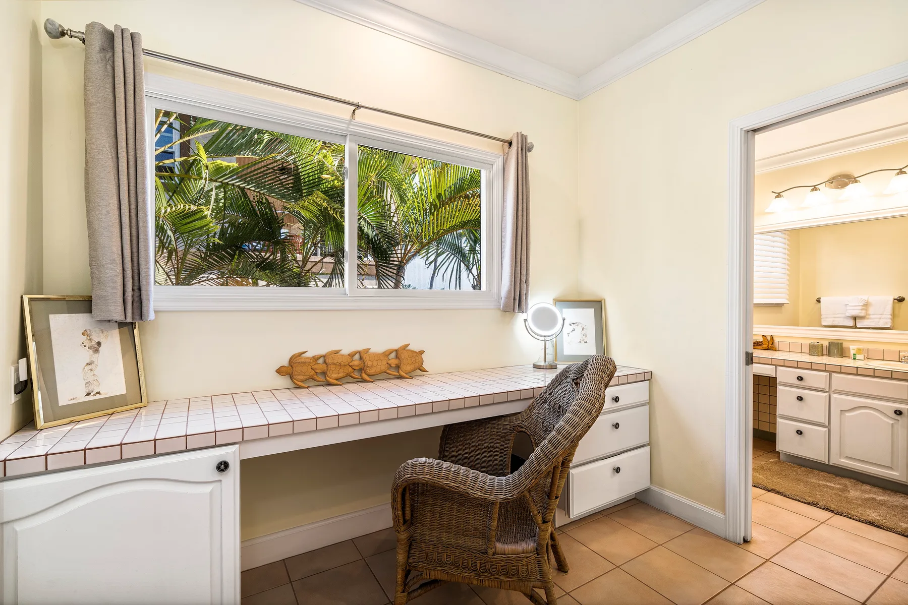 Un tocador de dormitorio con una silla de mimbre está bajo una ventana amplia con vista a palmeras.